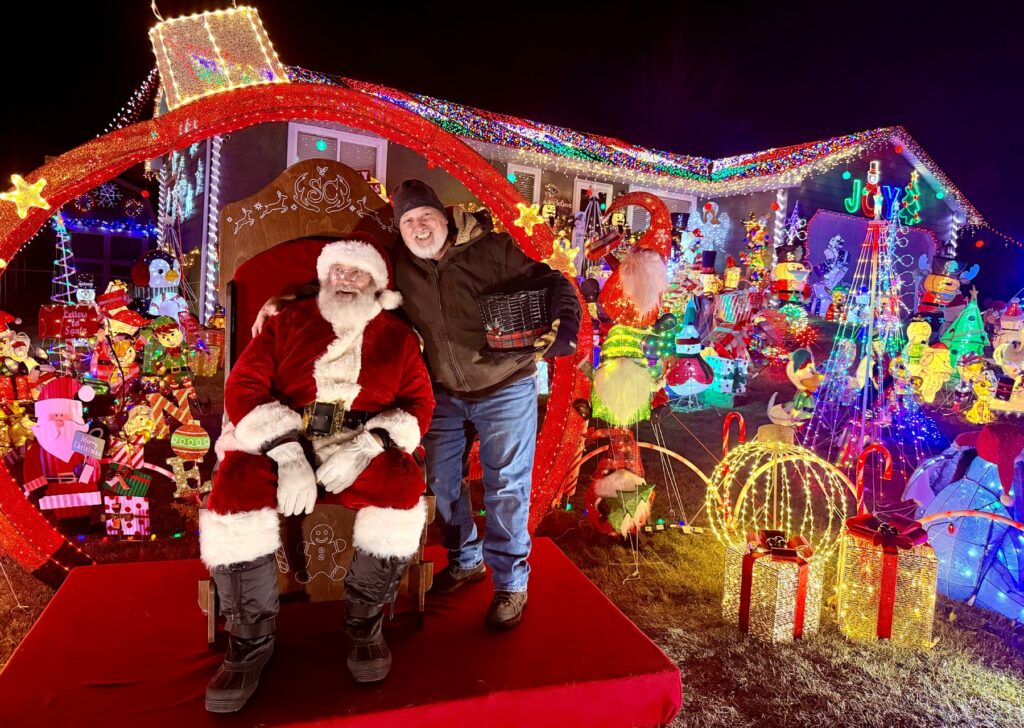 Dana Darger dressed as Santa beside a colorful Christmas light display.