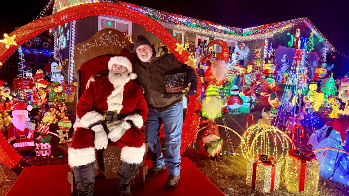 Dana Darger dressed as Santa beside a colorful Christmas light display.